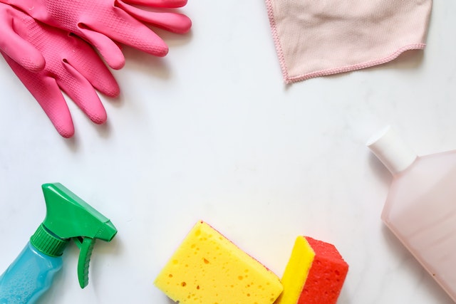 pink gloves, a cloth, a green spray bottle, yellow sponges, and a bottle of cleaning product on a white background